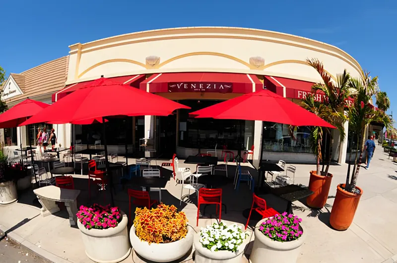 Venezia's storefront by day, crimson awnings and umbrellas over the sidewalk tables, flower planters in front